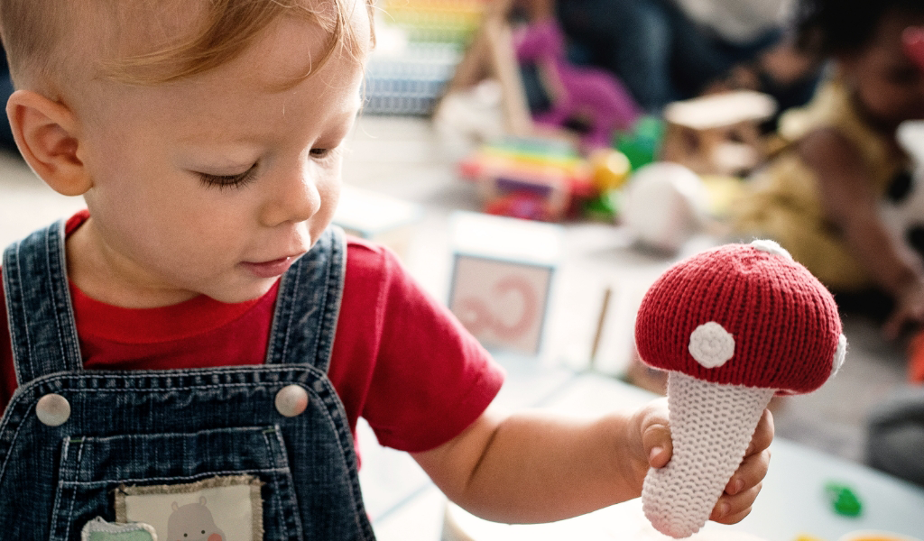 A young child at an Early Childhood Learning Center, dressed in denim overalls, holds a knitted red and white mushroom toy while playing indoors.