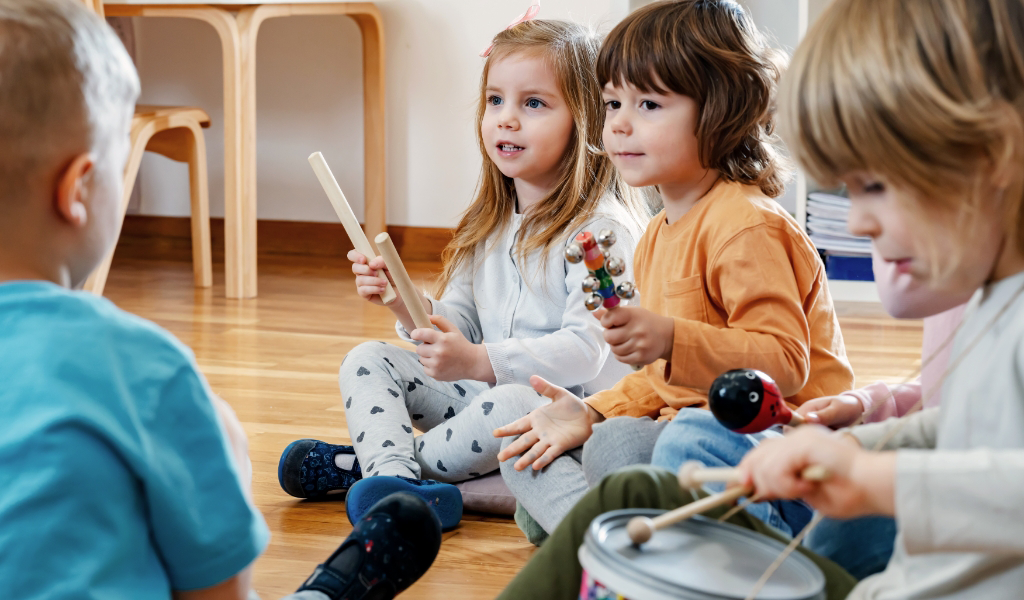 Four young children sit on the floor in an Early Childhood Learning Center classroom, playing various musical instruments including drumsticks, shakers, and a drum.