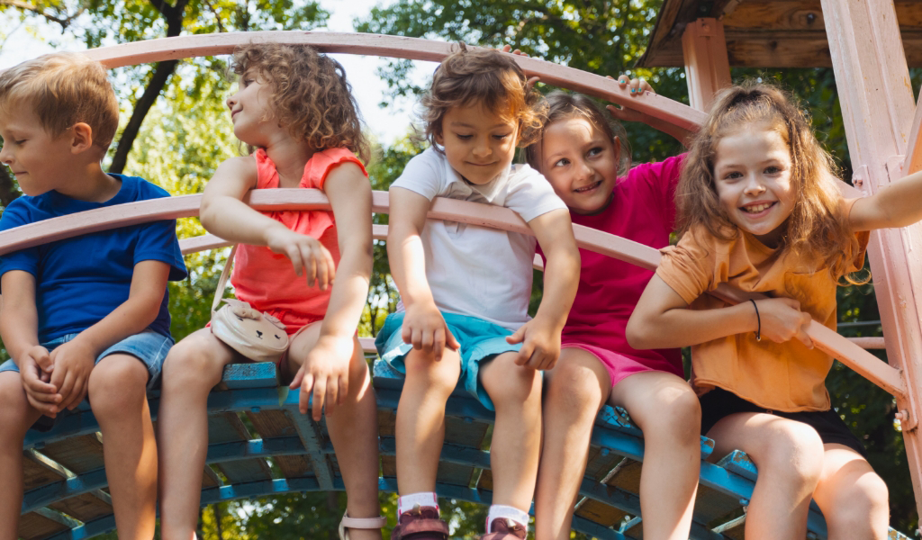 Five children from the Early Childhood Learning Center sit together on a playground structure outdoors, smiling and looking in different directions on a sunny day.