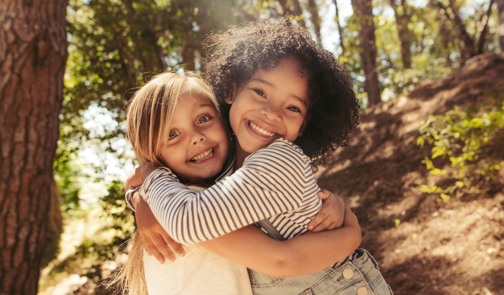 Two young children, from the Early Childhood Learning Center, are hugging and smiling outdoors in a sunlit forest area.