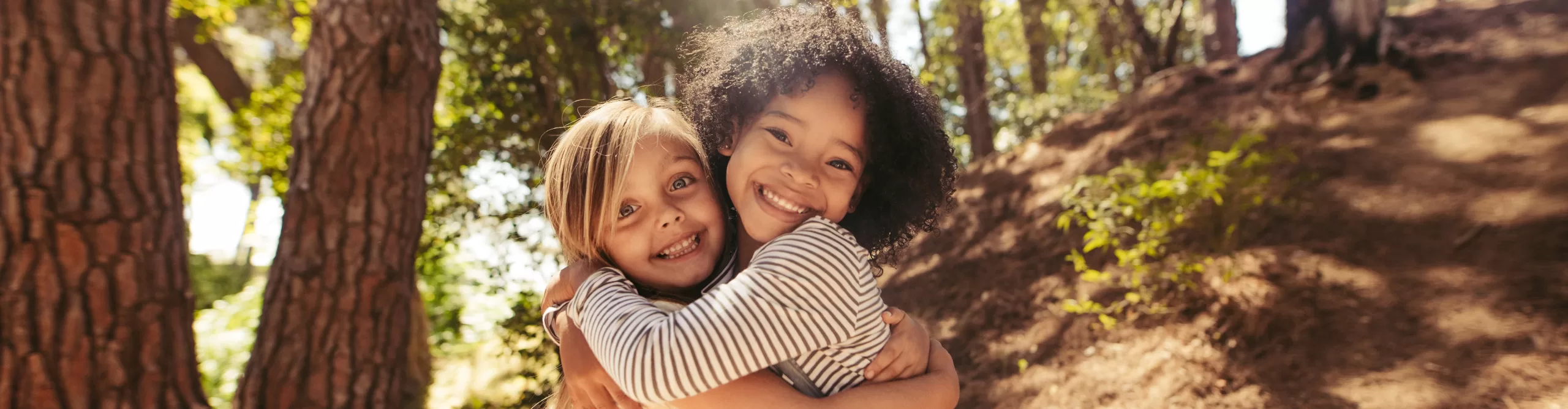 Two children smiling and hugging each other outdoors in a sunlit wooded area, enjoying the warmth and friendship fostered by After-School Care.