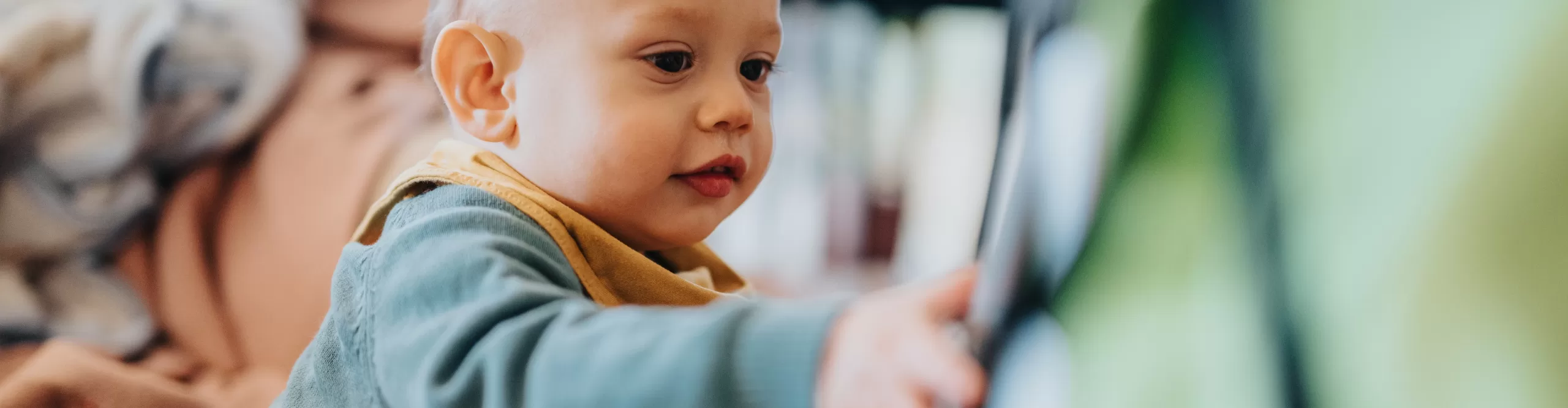 A baby wearing a green shirt and yellow bib reaches forward with interest, surrounded by a soft background of household items—perfectly capturing the wonder and curiosity often seen in an Infant Daycare setting.