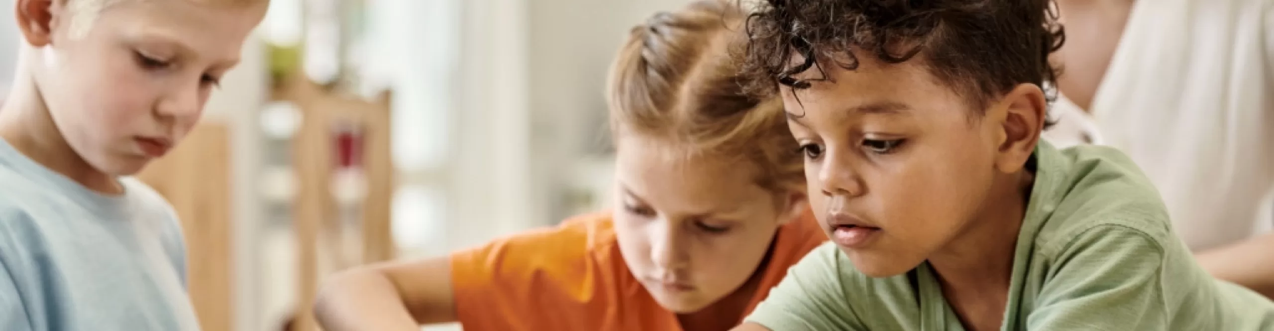 Three young children sit at a table, focused on an activity or project, while an adult from Pre-K School Care is partially visible in the background.