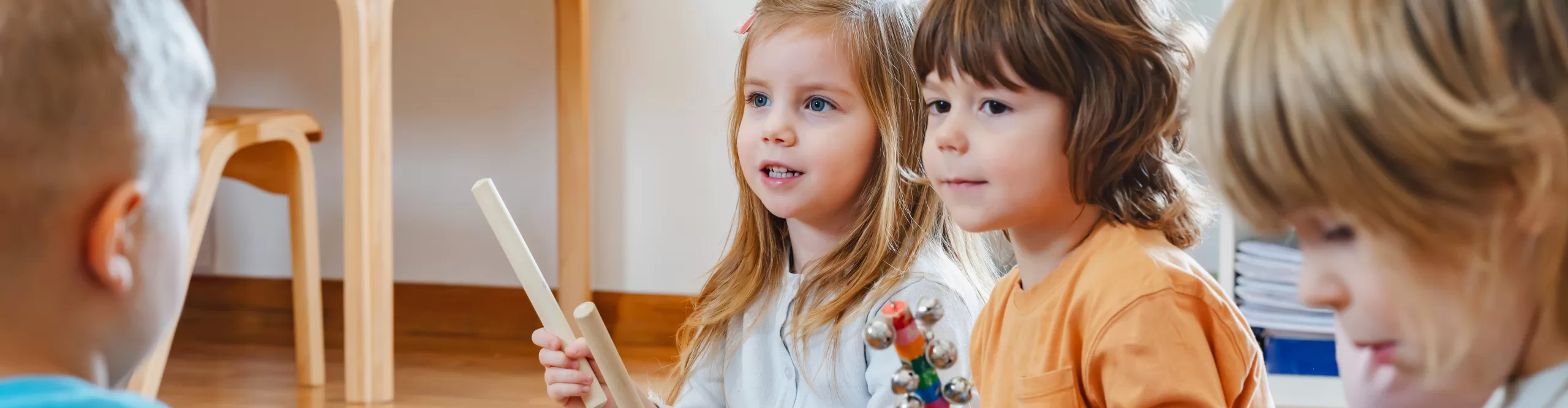 Several young children sit on the floor indoors during a Preschool Program, holding musical instruments and looking in the same direction.