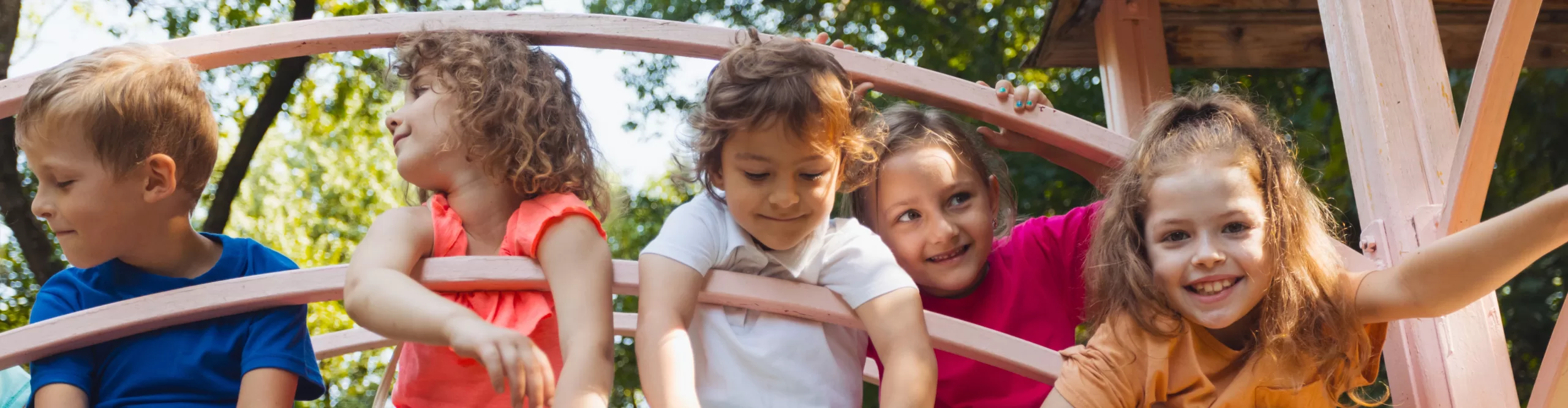 Five children sit and lean on a pink wooden structure outdoors at daycare summer camp, smiling and looking in different directions, with trees visible in the background.
