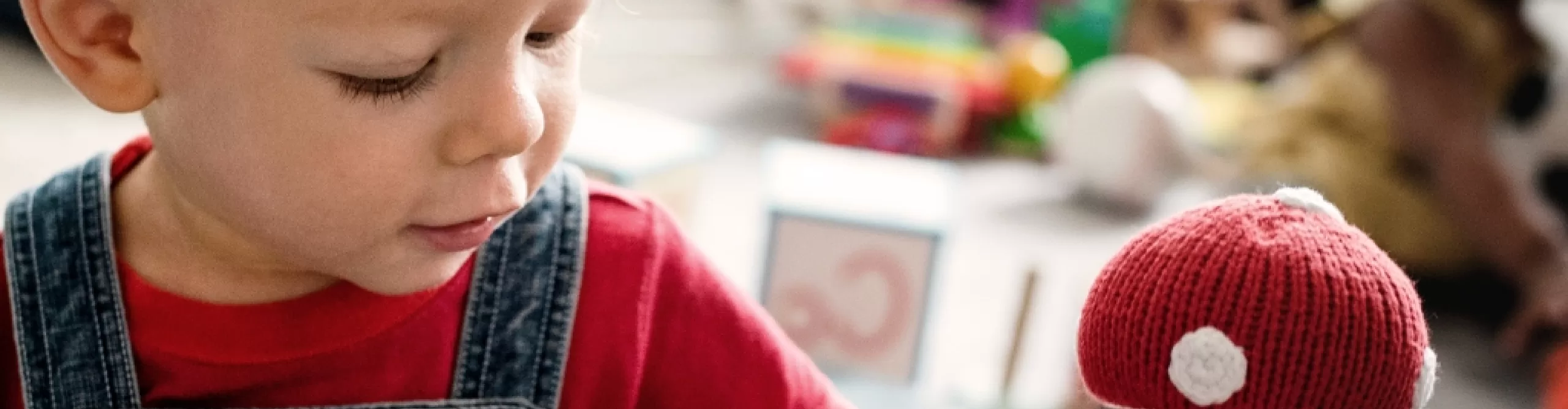 A young child in a red shirt and denim overalls at toddler daycare gazes at a red and white knitted toy, with other toys softly blurred in the background.