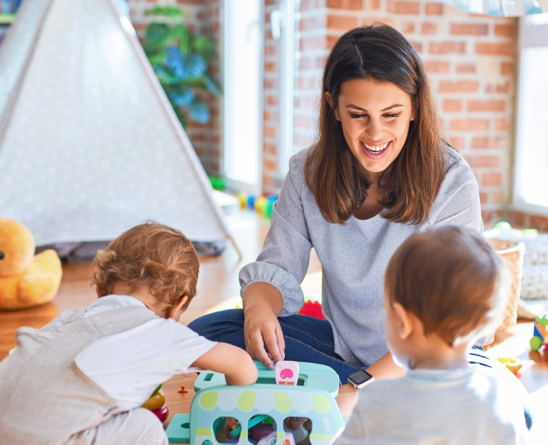 A woman sits on the floor indoors, smiling and playing with two young children and colorful toys—a joyful moment that reflects the heart of Childcare Careers.