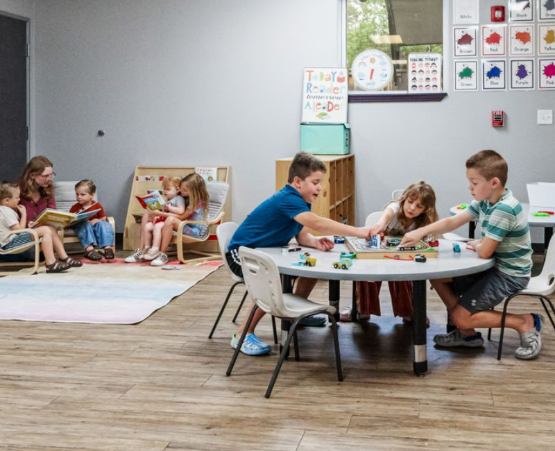 Children play with toys at a round table while others read books with adults on the left side of the Early Childhood Learning Center classroom.