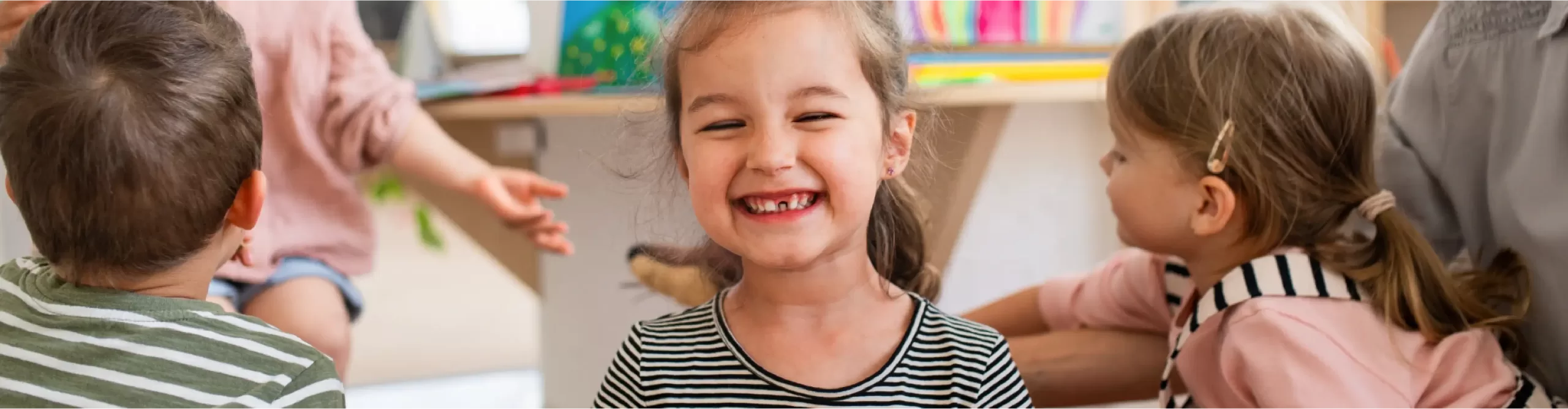 A young girl smiles at the camera while sitting among other children in a brightly lit indoor setting.
