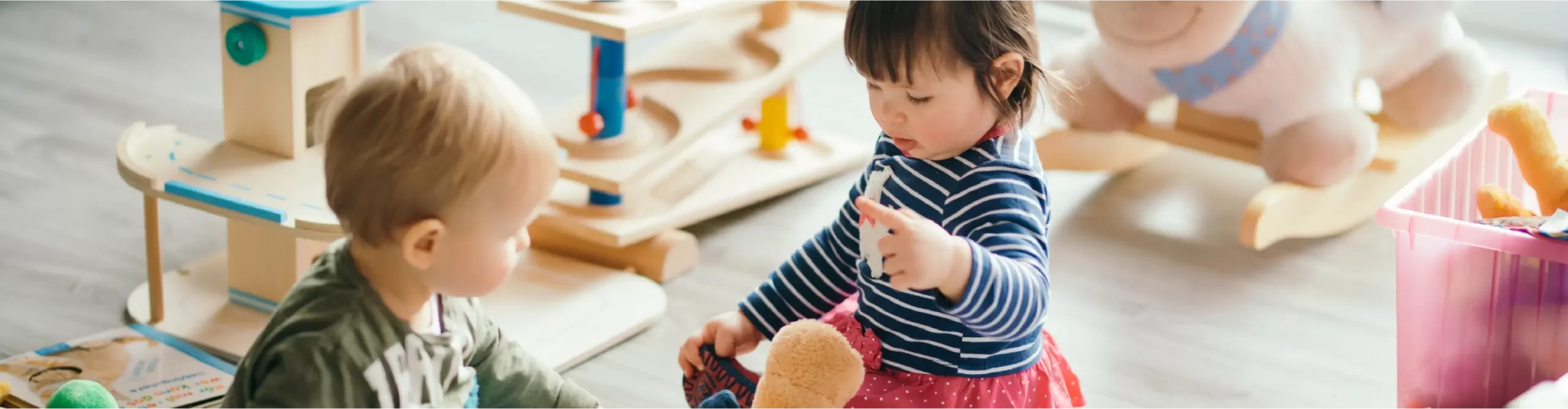 Two young children sit on the floor playing with toys in a brightly lit room with various wooden and plush toys around them.