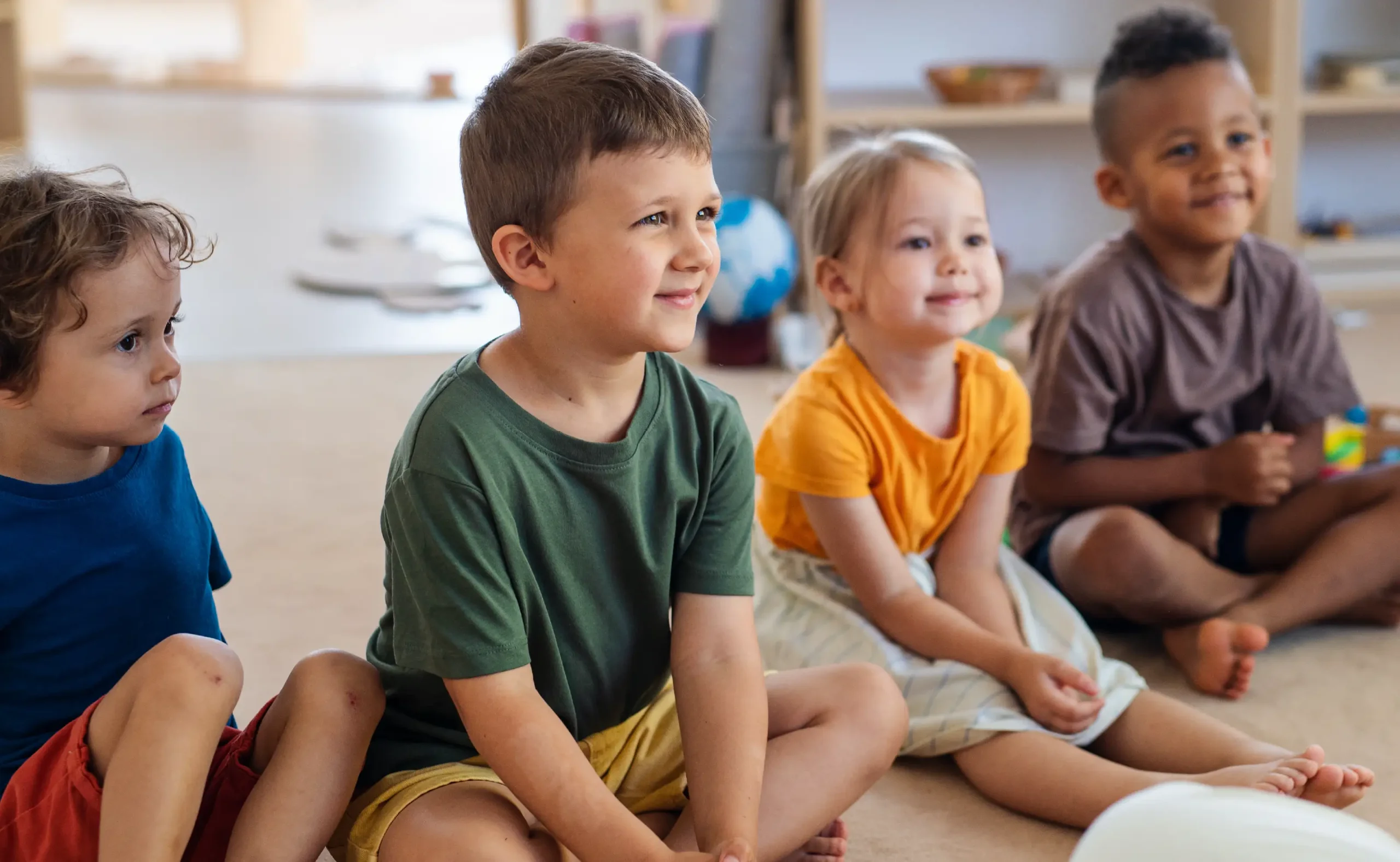 Group of small smiling children sitting on floor indoors in classroom.